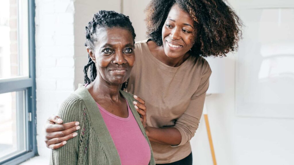 black woman caring for her elderly mother, hands on her shoulders; she smiles at her